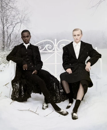Two fashion models in black outfits pose on a white bench in a snowy outdoor setting with bare trees and soft, diffused lighting.