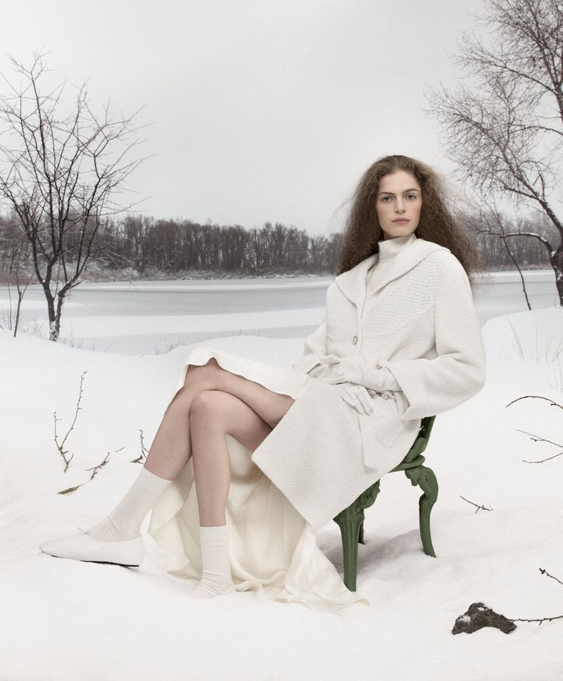 Woman in white coat sitting on green chair in snowy winter landscape, wearing white gloves, socks, and shoes. Winter fashion photo shoot.