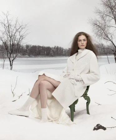 Woman in white coat sitting on green chair in snowy winter landscape, wearing white gloves, socks, and shoes. Winter fashion photo shoot.