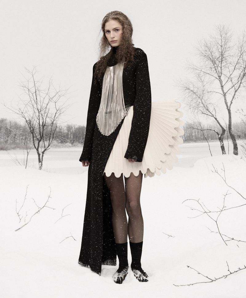 A fashion model poses in a snow-covered landscape, wearing a black coat with an asymmetrical skirt, shiny silver top, and black stockings.
