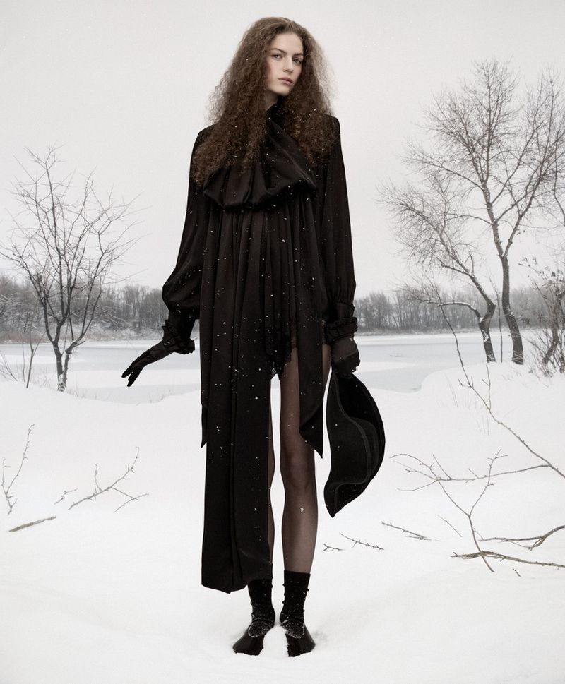 Fashion shot of an adult female in a black dress and gloves, posing in a snowy outdoor environment with bare trees and an overcast sky.