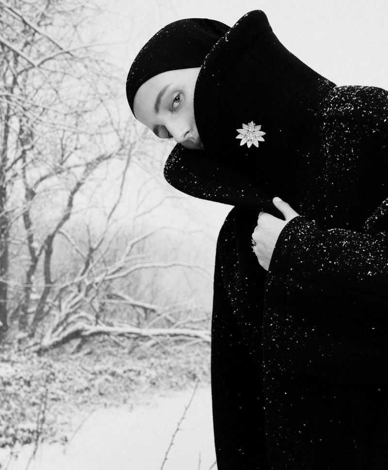 A person wearing a black coat with a brooch and a matching hat stands in a snowy outdoor environment, in a black and white aesthetic.