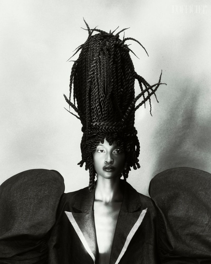 Monochrome portrait of a Black female model wearing a dark blazer and an elaborate braided updo hairstyle against a grey background.