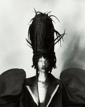 Monochrome portrait of a Black female model wearing a dark blazer and an elaborate braided updo hairstyle against a grey background.