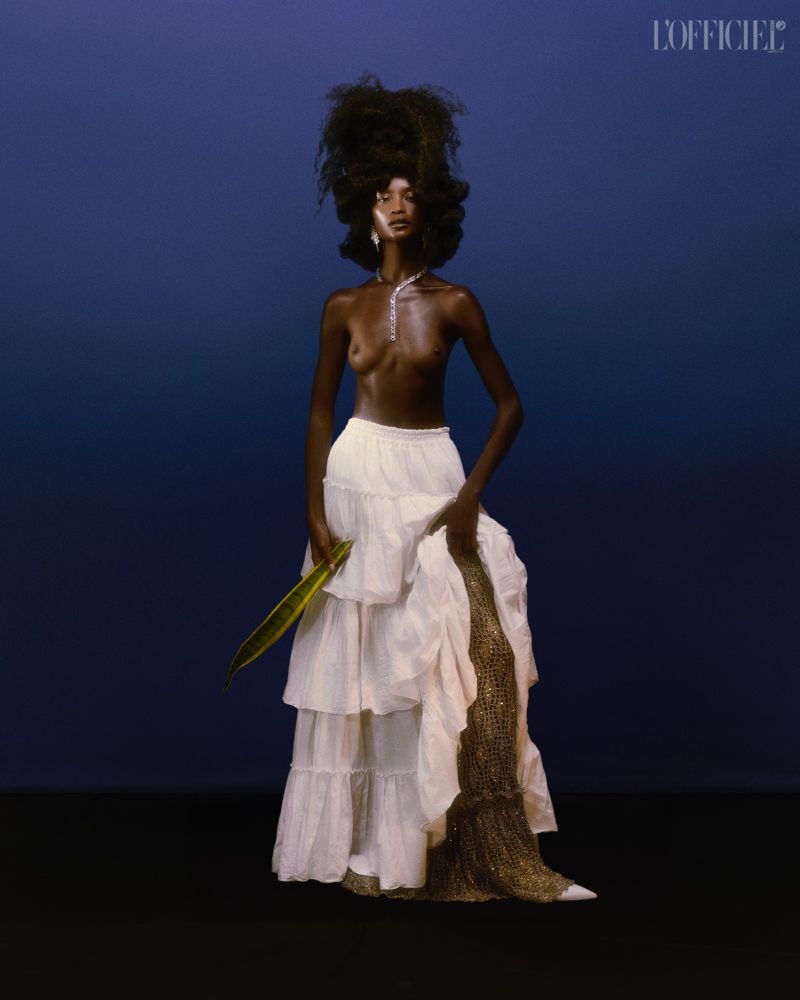 Dark-skinned woman with elaborate afro hairstyle posing in a photo studio wearing a white ruffled skirt and a snake plant.