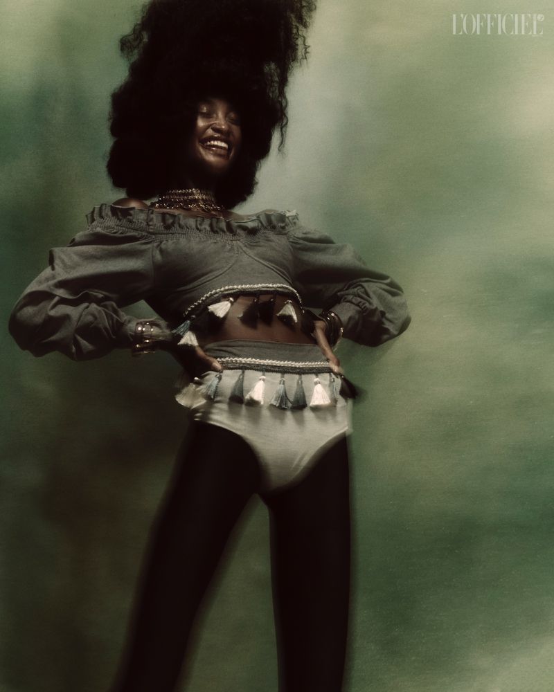 Portrait of smiling Black woman with afro in off-shoulder top and bottoms with tassels, posing against a blurred green backdrop.