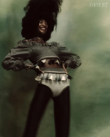 Portrait of smiling Black woman with afro in off-shoulder top and bottoms with tassels, posing against a blurred green backdrop.