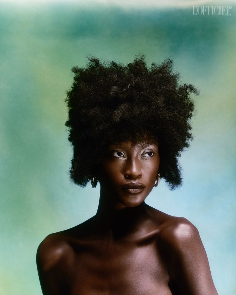Close-up portrait of a black woman with natural afro hair and hoop earrings set against a softly textured green and blue background.