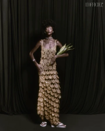 A model poses in a full-length, open-weave, fringed gold gown against a dark draped backdrop, holding a long green plant.