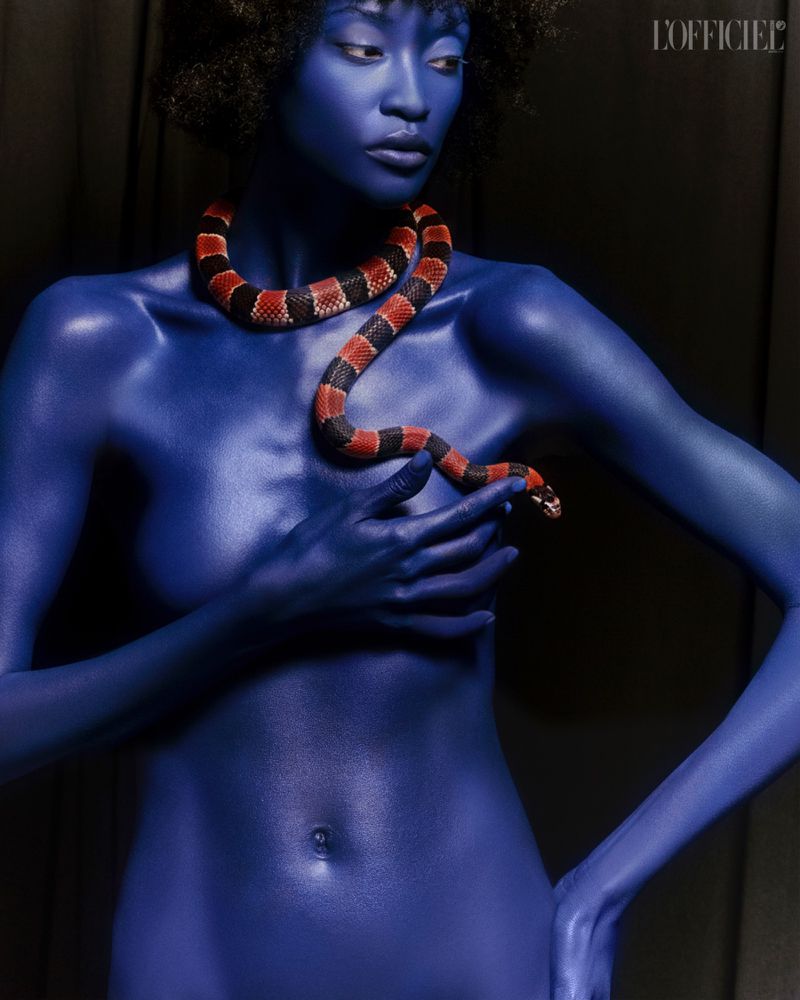 Striking fashion portrait of a woman with blue body paint, adorned with a snake, against a dark backdrop, showcasing a unique style.