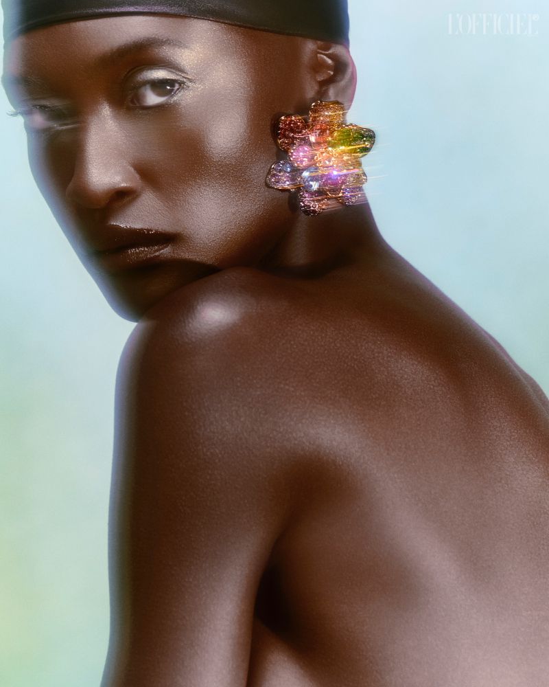 Close-up portrait of a bald, dark-skinned woman wearing a black head covering and iridescent, multi-colored flower earrings.