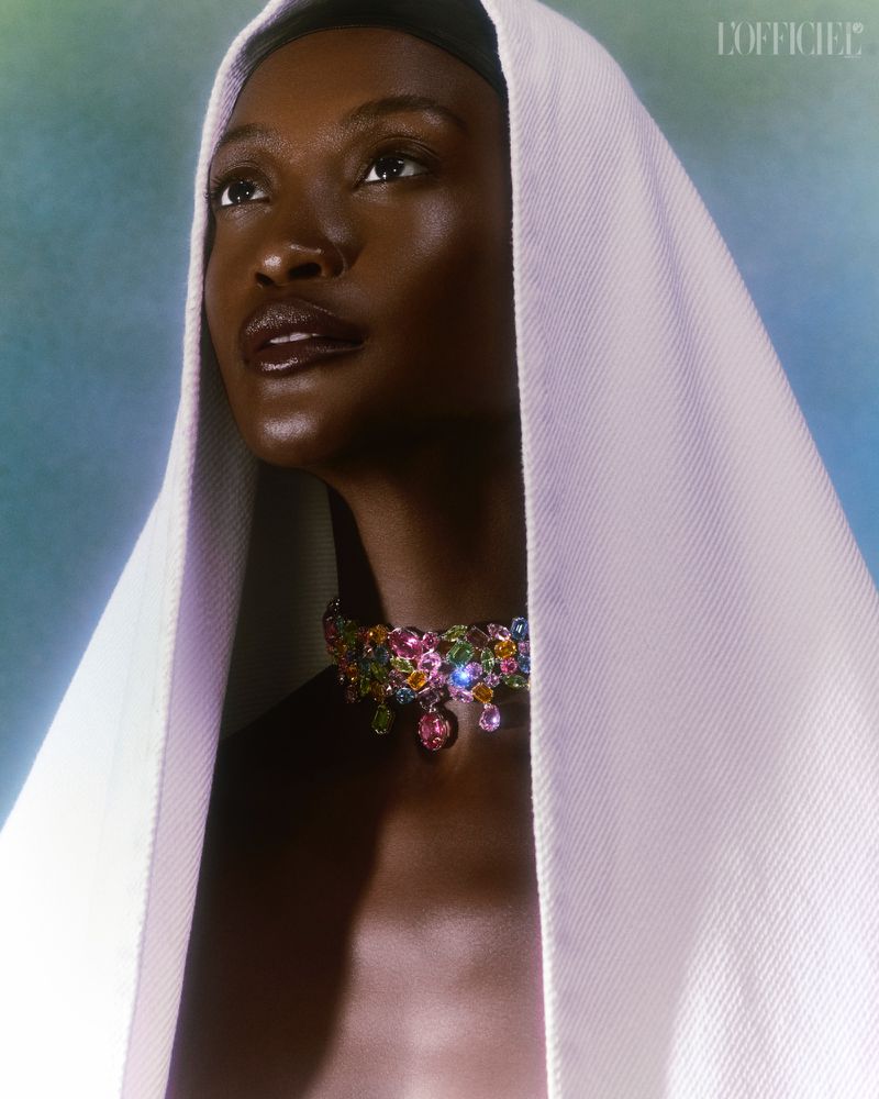 Close-up portrait of a Black woman wearing a white headscarf and colorful jeweled necklace, gazing upwards in a studio setting.