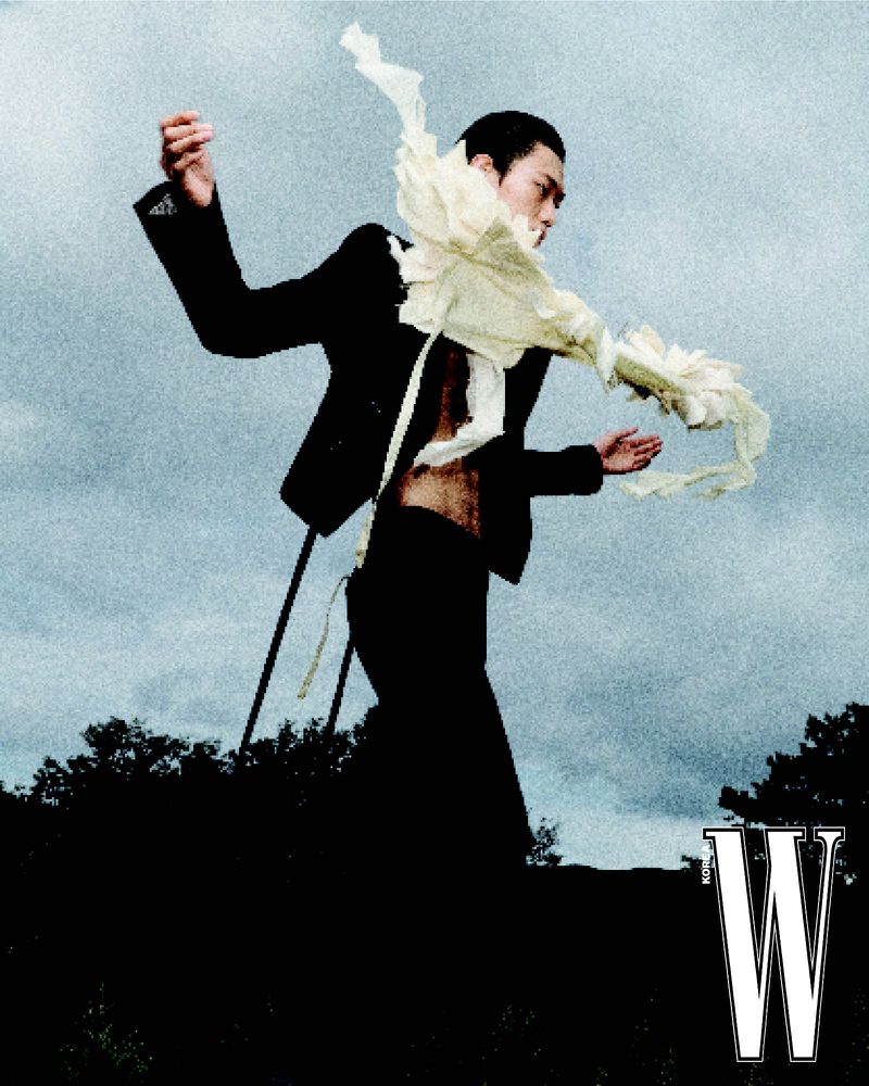 Male fashion model in black suit with dramatic white frill poses outdoors with blue sky backdrop and W Magazine logo.