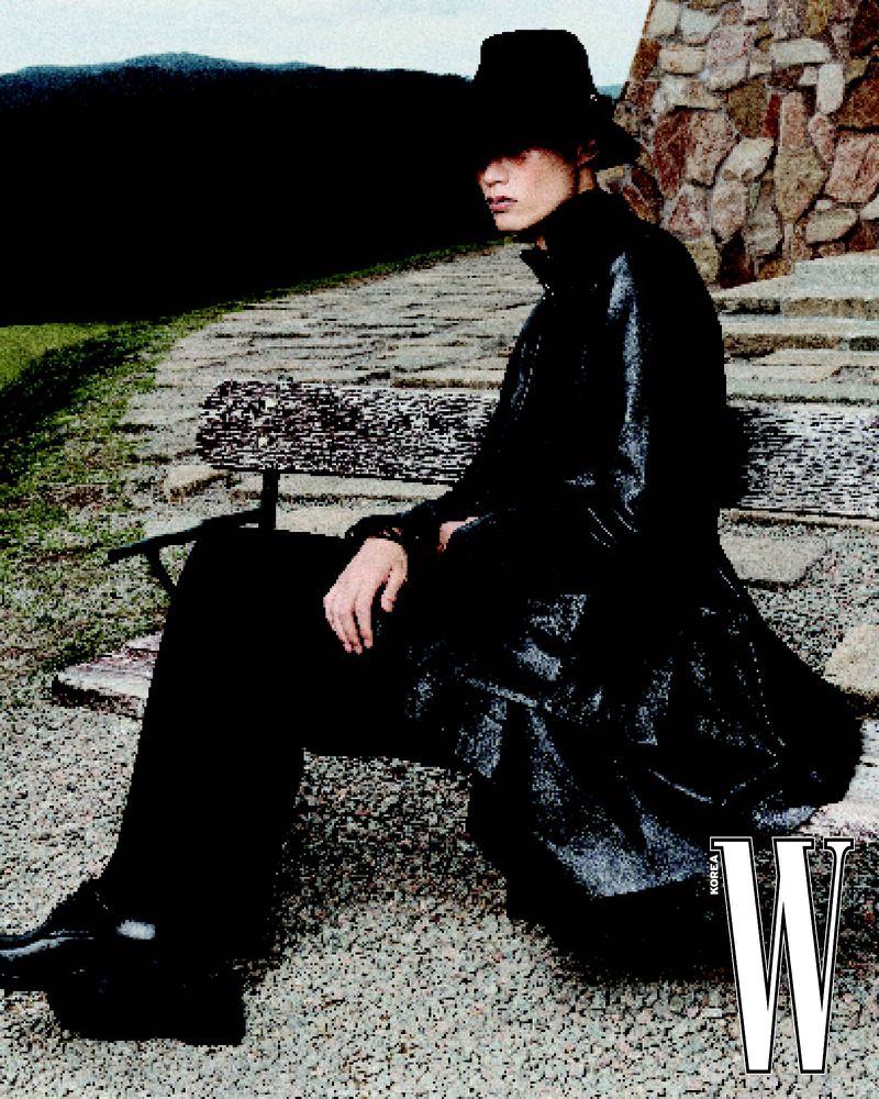 A stylish person in a hat and long coat sits on a stone bench, accented by stonework and natural scenery, with a bold 'W' logo.