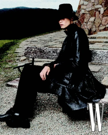 A stylish person in a hat and long coat sits on a stone bench, accented by stonework and natural scenery, with a bold 'W' logo.
