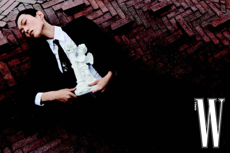 An adult male model in a black suit and white shirt, holds an angel sculpture while lying on a brick surface, shot for W Korea.