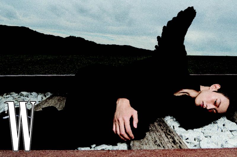 Fashion editorial shot of an adult male model lying on railroad tracks in a black outfit, with a mountain background under a cloudy sky.