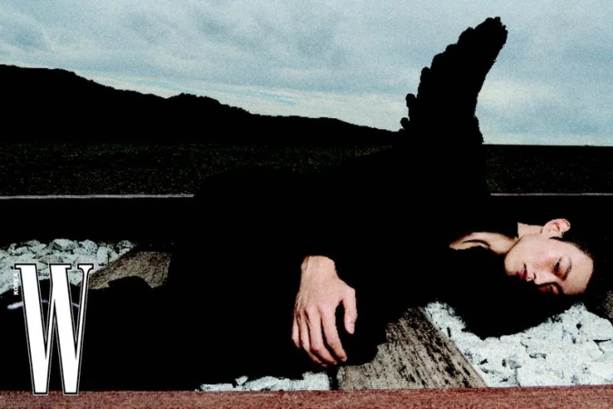 Fashion editorial shot of an adult male model lying on railroad tracks in a black outfit, with a mountain background under a cloudy sky.