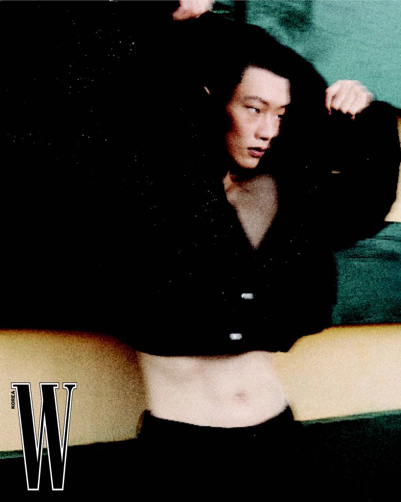 Portrait of a young Asian male model wearing a dark crop top with buttons, posing against a multi-colored geometric background indoors.