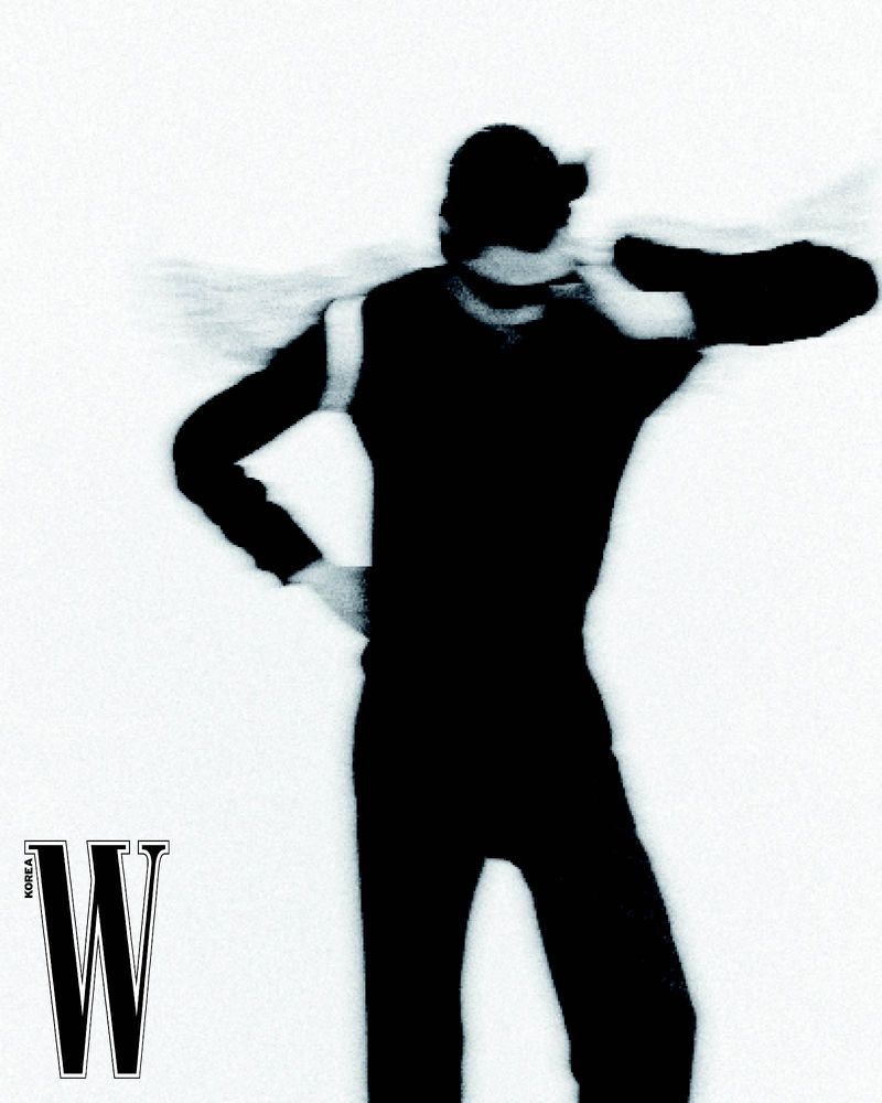Back view of silhouetted figure in dark clothing with motion blur against a white background. W Korea logo visible.