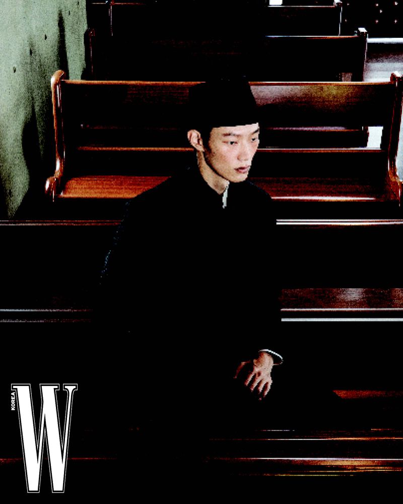 Young Asian man in a black shirt and cap sitting in a wooden church pew looking down with magazine logo in the lower left corner.