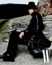 Model Hoyong Kim wearing a wide-brim black hat and long black coat, seated on a rustic wooden bench on an outdoor path.