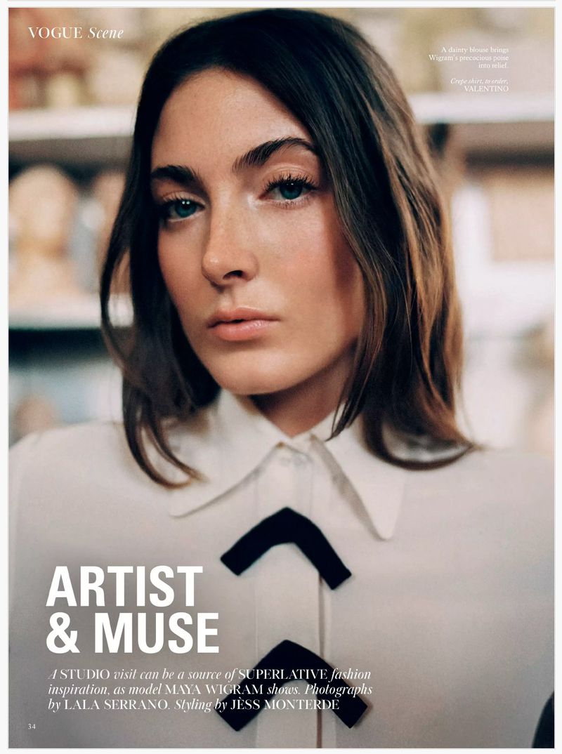 Close-up editorial fashion shot of a young adult woman with long dark hair, wearing a white blouse with black bow details, indoors.