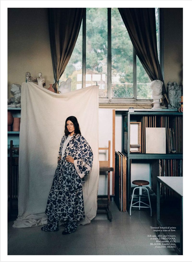 Woman in floral robe stands in art studio near canvases and sculptures with natural light streaming through large window.
