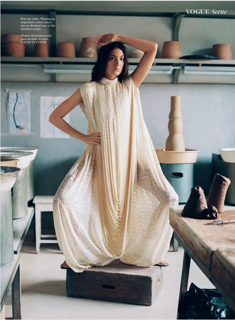 A woman poses in a pale yellow dress in a ceramics studio, standing on a wooden box, surrounded by pottery and tools.