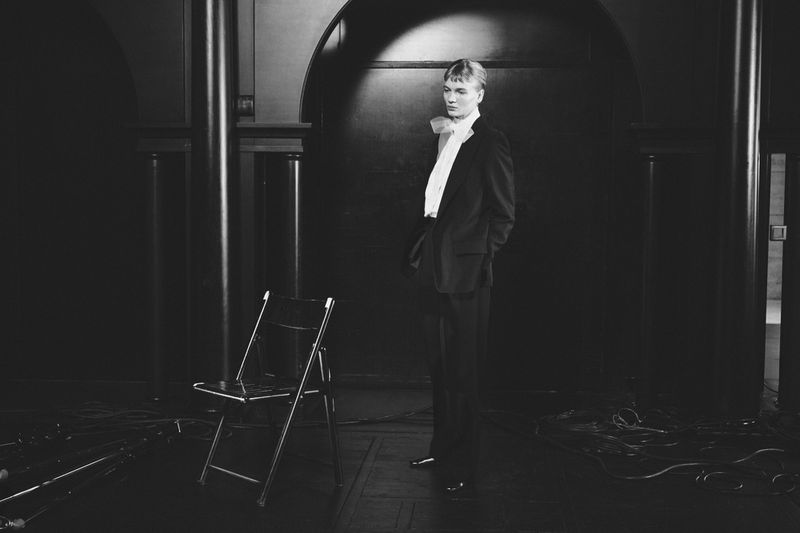 A young adult male model poses in a dark room wearing a suit and bow. An empty metal folding chair sits nearby in the shadows.