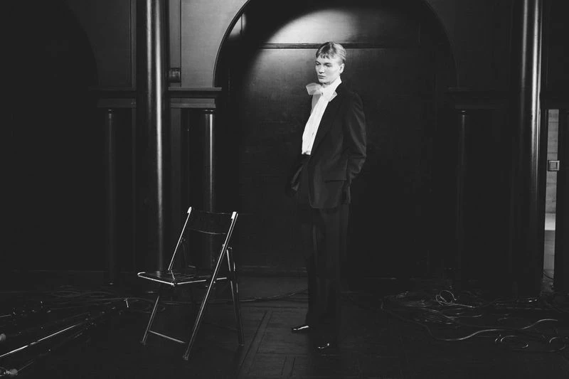 A young adult male model poses in a dark room wearing a suit and bow. An empty metal folding chair sits nearby in the shadows.