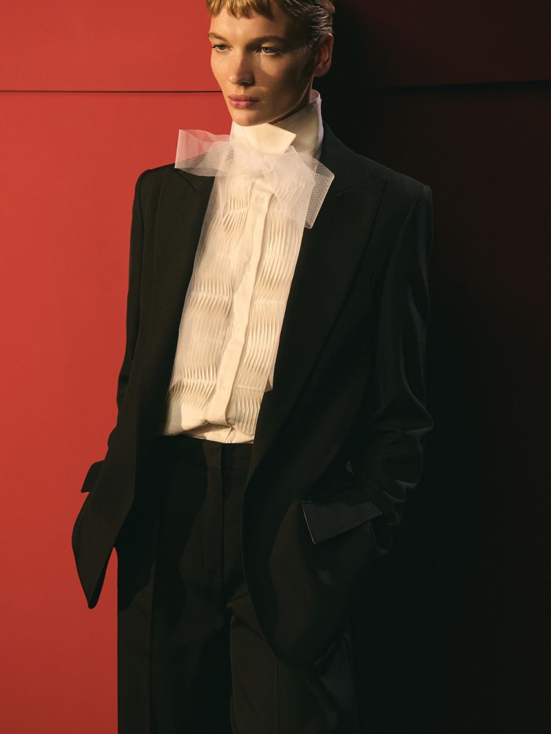 Fashion portrait of a woman wearing a black suit and white pleated shirt with a bow, against a gradient red and black background.