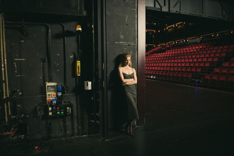 An adult woman leans against a wall on stage, wearing a dark dress, while the red seating of the theatre is in the background.