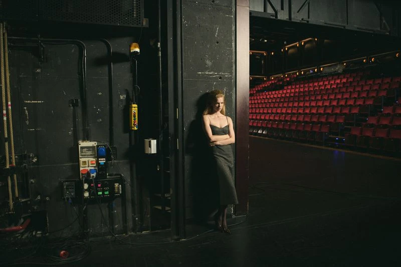 An adult woman leans against a wall on stage, wearing a dark dress, while the red seating of the theatre is in the background.
