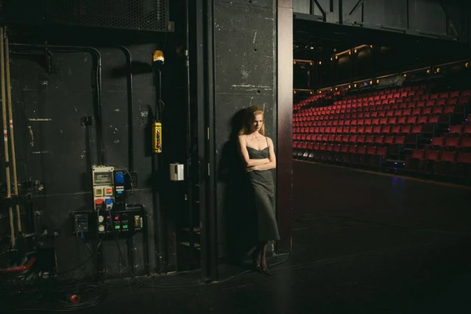 An adult woman leans against a wall on stage, wearing a dark dress, while the red seating of the theatre is in the background.