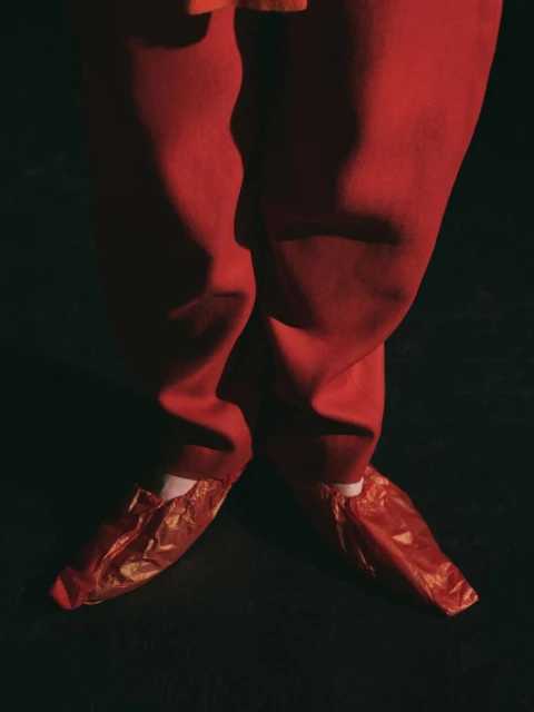 Close-up of human legs and feet wearing red trousers and red shoe covers, set against a dark backdrop in a studio photograph.