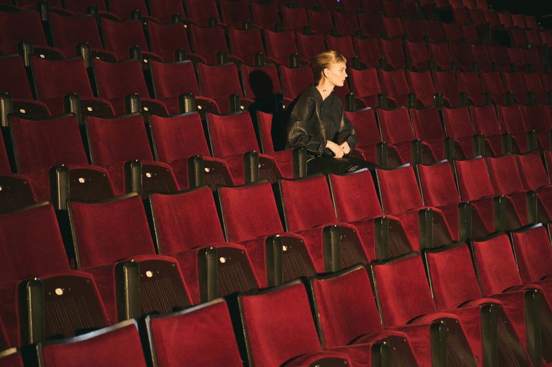 Woman in black leather jacket sits alone in a red velvet seat inside a dark theatre with rows of empty seats in the background.