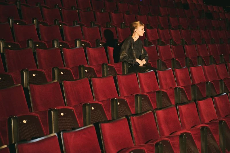 Woman in black leather jacket sits alone in a red velvet seat inside a dark theatre with rows of empty seats in the background.