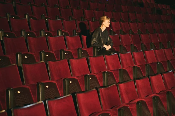 Woman in black leather jacket sits alone in a red velvet seat inside a dark theatre with rows of empty seats in the background.