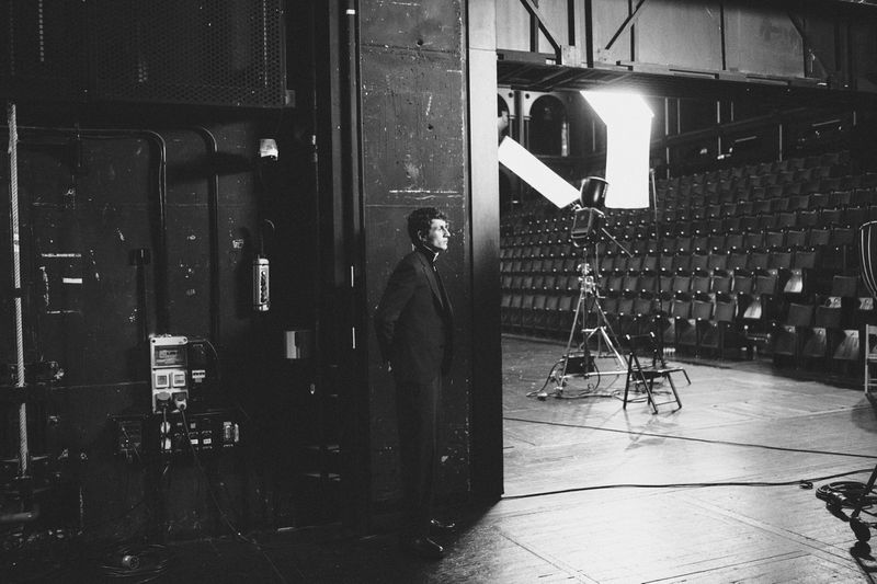 Black and white shot of a man in suit standing on a stage near theatrical lighting equipment, with rows of empty chairs in the background.