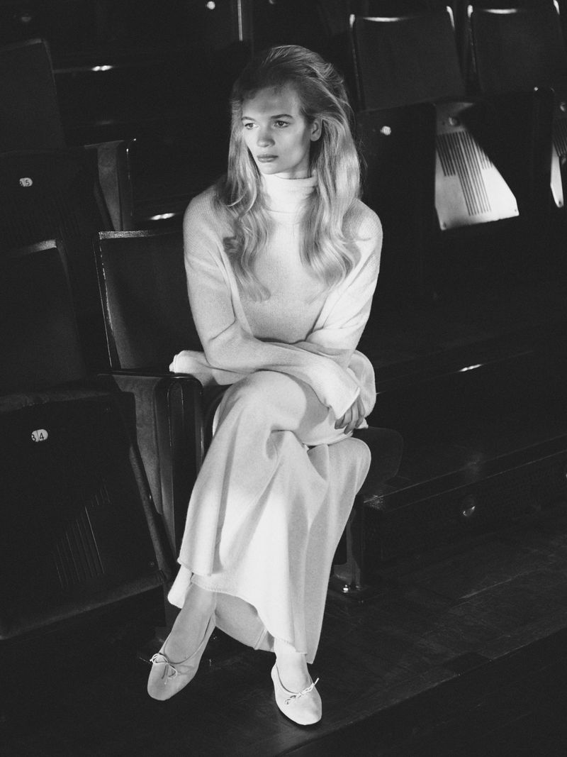 Woman in a white turtleneck and long dress sits in a theatre seat. She is wearing flats, and the scene is grayscale.