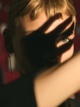 Close-up portrait of a woman with short bangs and a dangling earring, with a sharp hand shadow cast across her eyes and face.