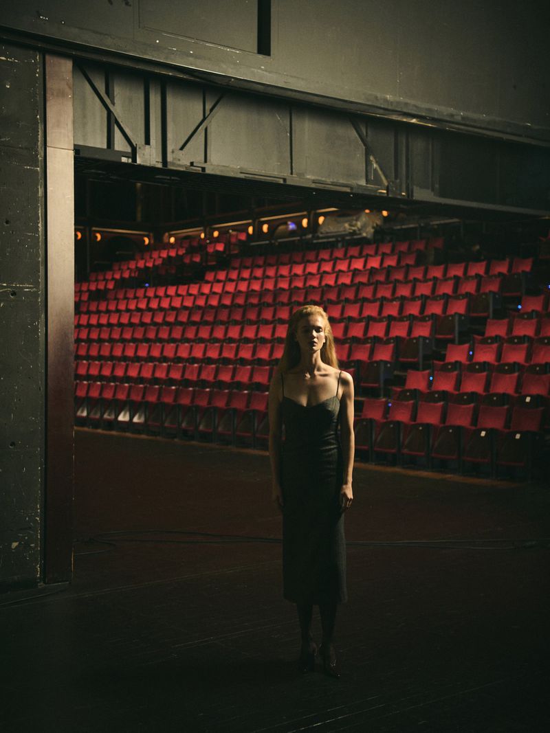 A woman in a dark dress stands on a theatre stage with rows of red seats in the background, bathed in dramatic lighting.