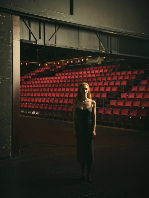 A woman in a dark dress stands on a theatre stage with rows of red seats in the background, bathed in dramatic lighting.