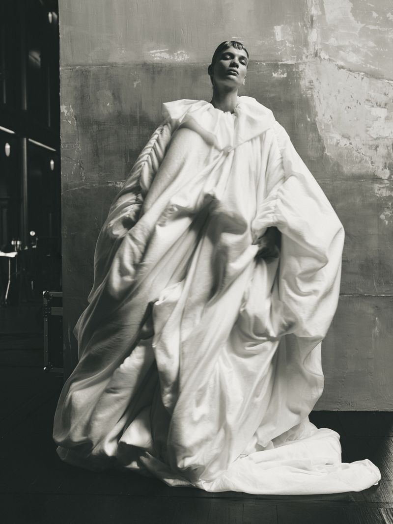 Fashion model in an oversized white garment poses in an indoor setting with textured walls and subdued lighting, capturing an editorial style.