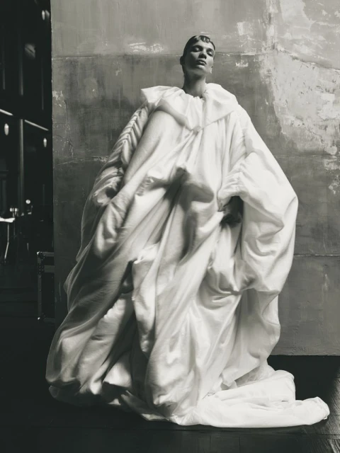Fashion model in an oversized white garment poses in an indoor setting with textured walls and subdued lighting, capturing an editorial style.