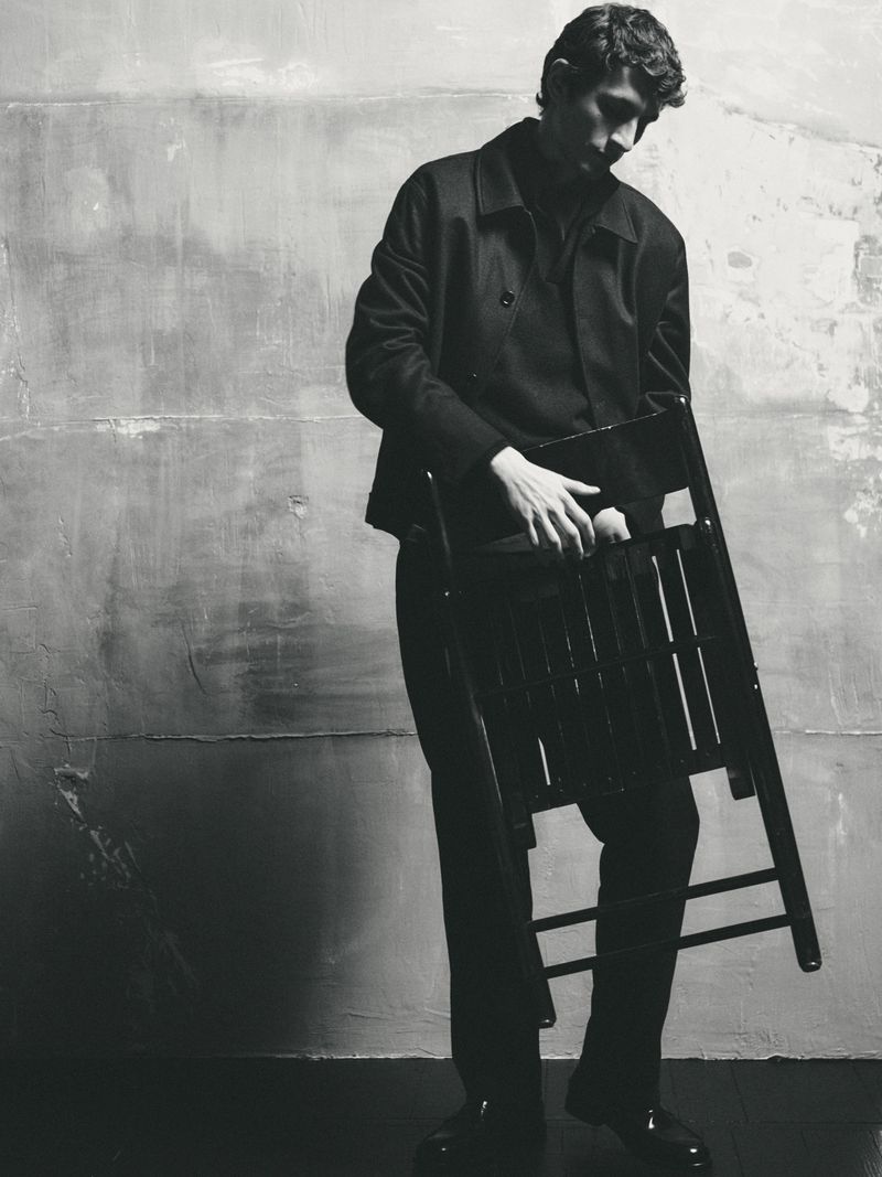 Monochrome portrait of a man holding a folding chair in front of a textured wall in an indoor studio setting.