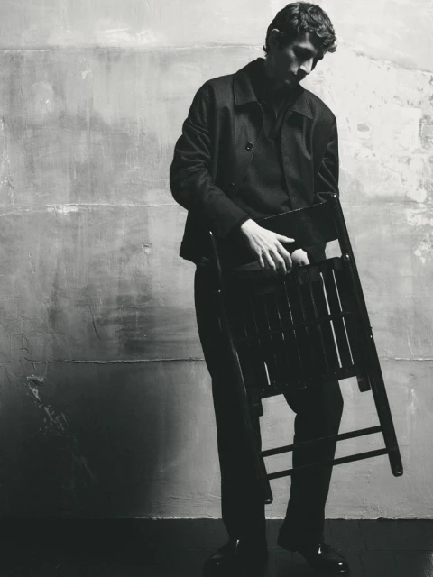 Monochrome portrait of a man holding a folding chair in front of a textured wall in an indoor studio setting.