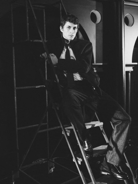 Monochrome portrait of a young man in a suit, seated on a metal ladder against a backdrop with geometric shapes.