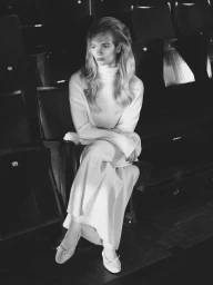 Model Caitlin Soetendal seated in an empty theater chair wearing a long white knit turtleneck dress and ballet flats in black and white.
